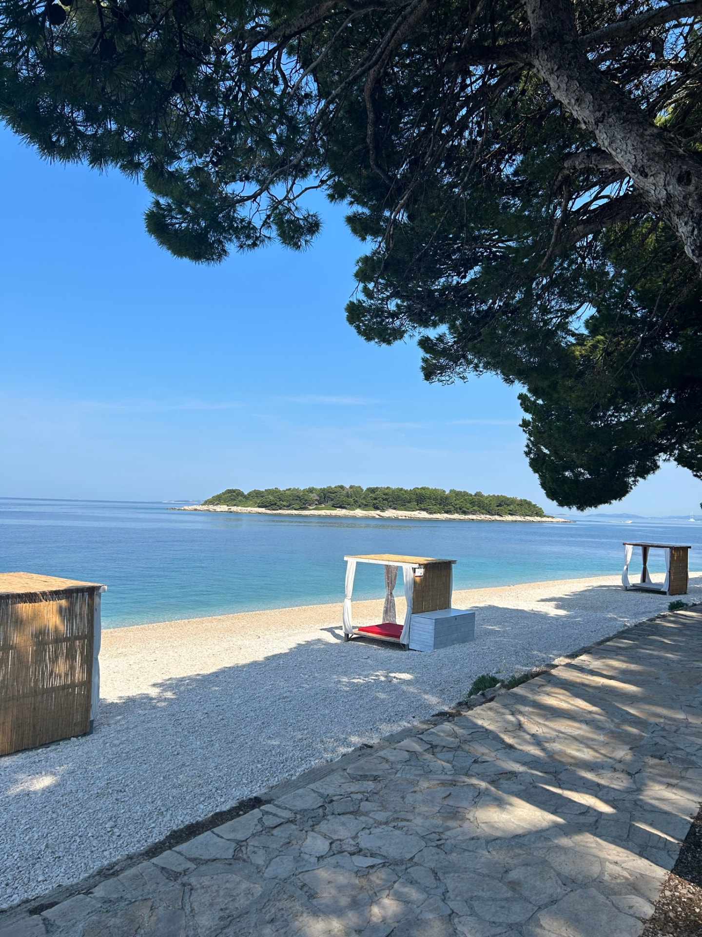 Pebble beach with cabanas at the tip of the Raduca peninsula, seen from under a pine tree